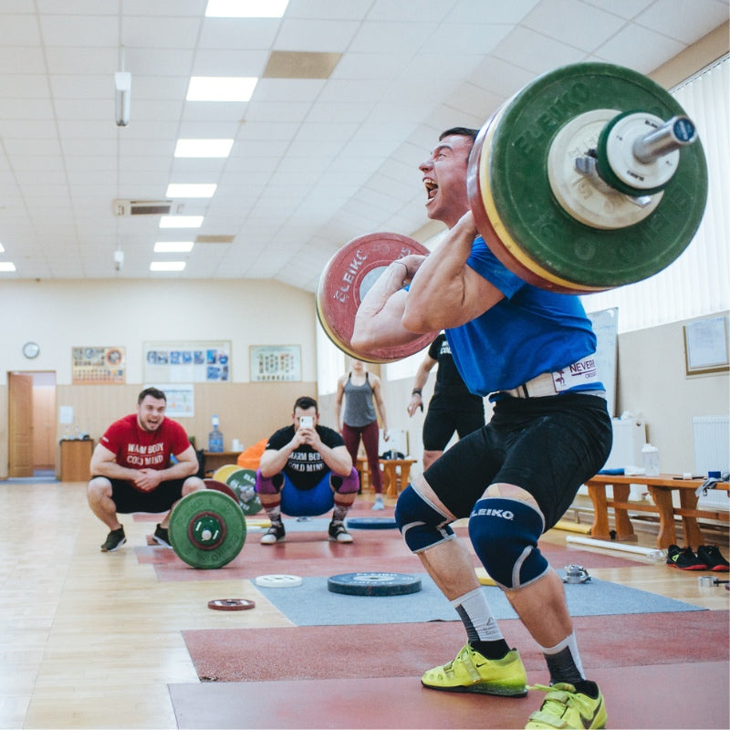 Person lifting a barbell in a weightlifting gym with other people in the background. (Torokhtiy Weightlifting)