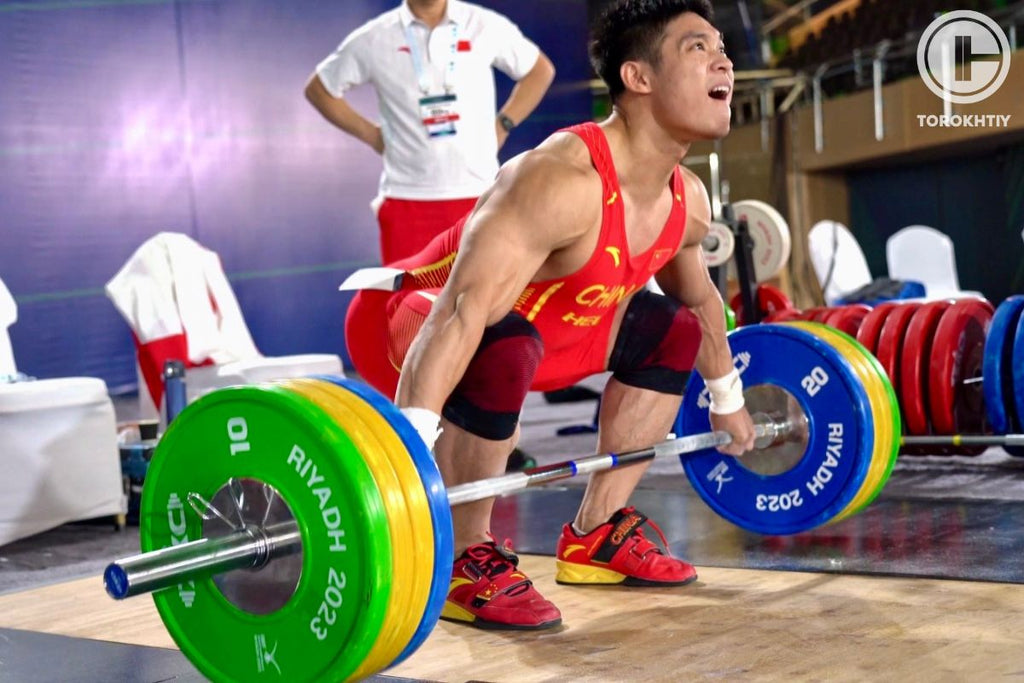 Weightlifter in a red and black outfit lifting a loaded barbell with 'Riyadh 2023' on it.