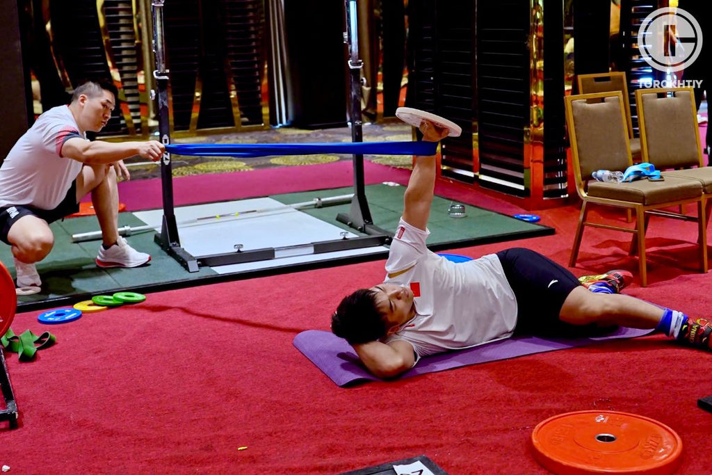 Person exercising on a mat in a gym setting with another person observing.