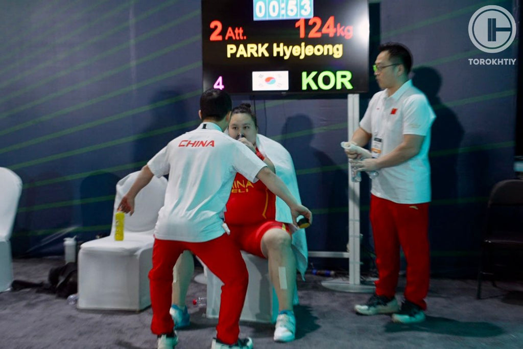 Athlete in red and white outfit being weighed by officials with a digital display in the background